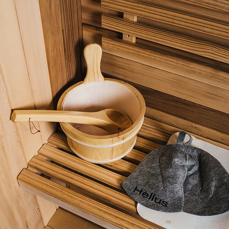 Wooden bucket and ladle on a wooden bench in a sauna with a towel and 'Helius' branding.